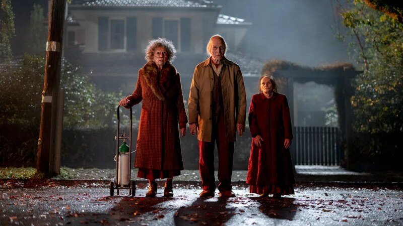 Three people stand on a wet street at night outside a house, with an oxygen cylinder beside them.