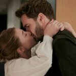 A woman in a white blouse kisses a man in a dark shirt, with her arms around his neck in a hallway.