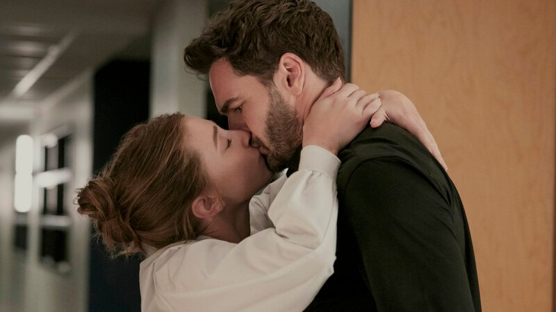 A woman in a white blouse kisses a man in a dark shirt, with her arms around his neck in a hallway.