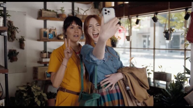 Two women pose for a selfie inside a plant-filled shop in an official Let’s Have Kids! trailer still.