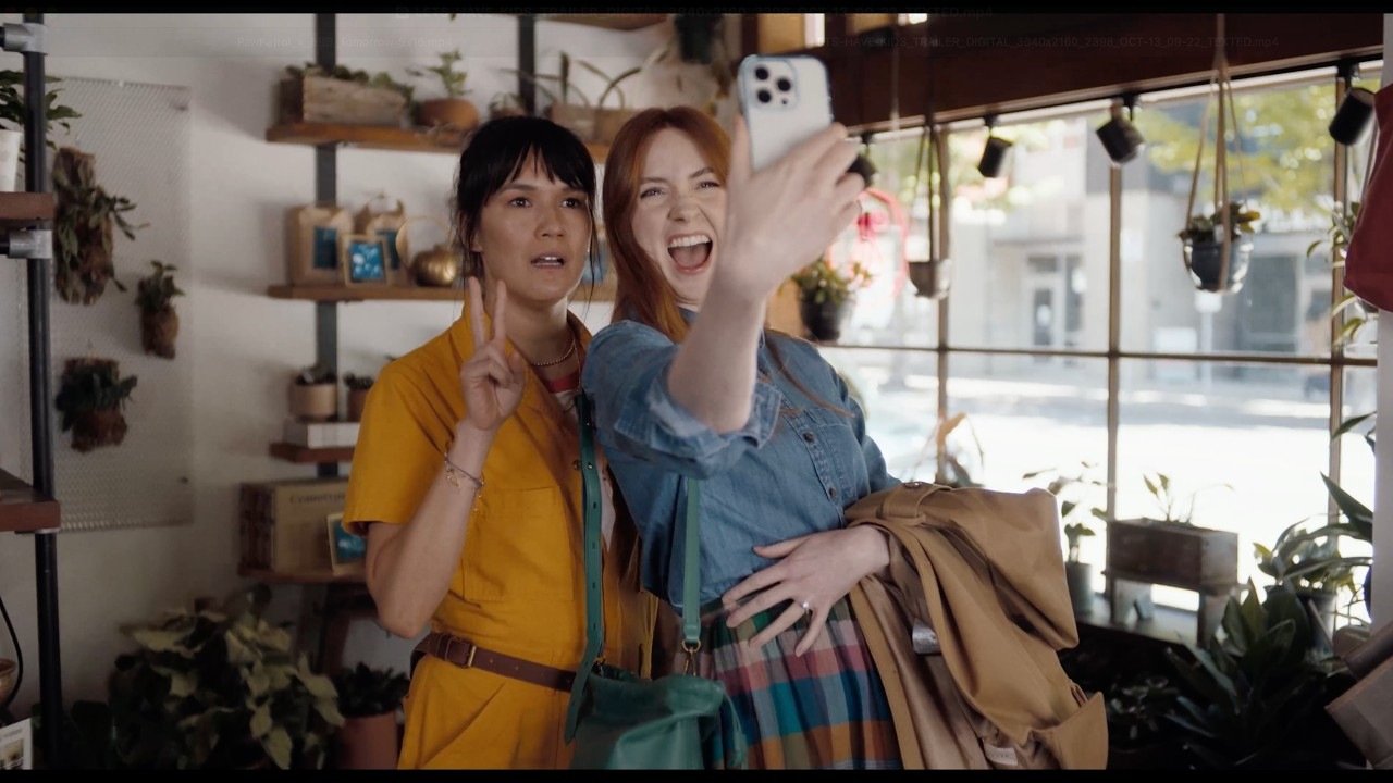 Two women pose for a selfie inside a plant-filled shop in an official Let’s Have Kids! trailer still.